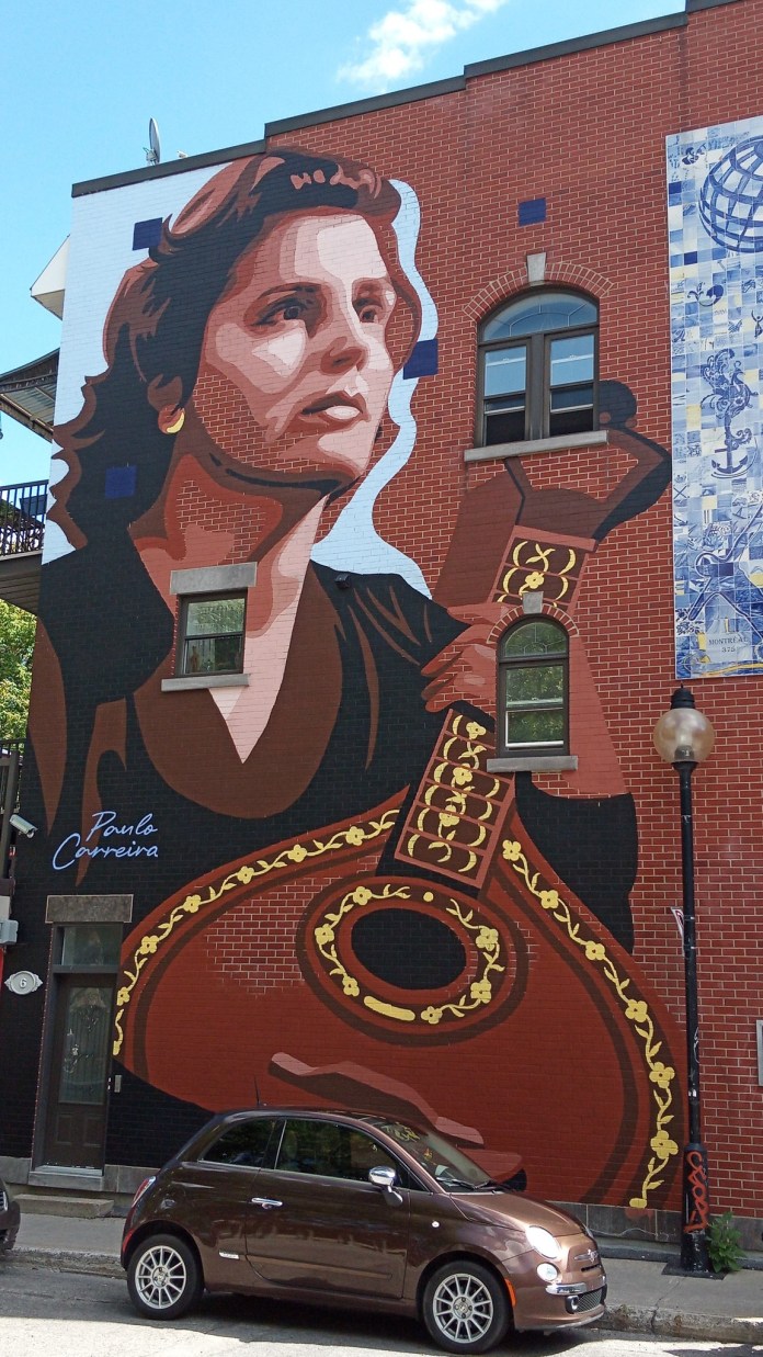 Bold, stylized mural of a woman holding a Portuguese guitar painted across the façade of a red brick building in Montreal with windows integrated into the artwork.