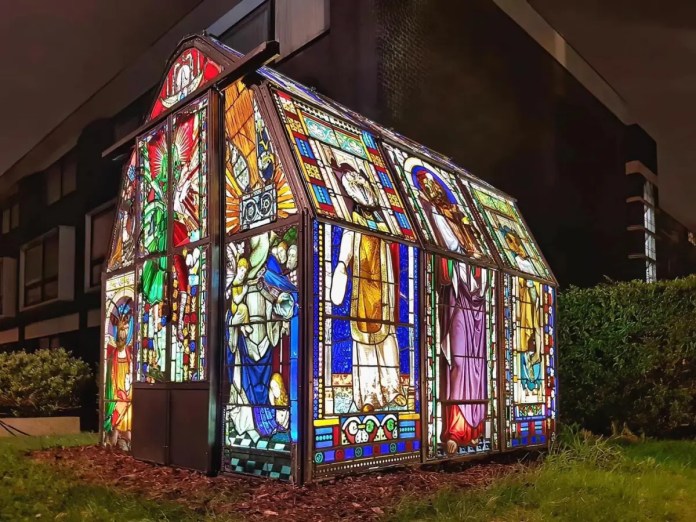 A stunning greenhouse made from repurposed stained glass windows from old churches, designed by artists Heywood and Condie in London. The structure glows brilliantly at night, showcasing intricate religious imagery, saints, and decorative patterns in vibrant blues, reds, and yellows. Surrounded by modern buildings, this unique installation contrasts history with contemporary urban life, blending art, sustainability, and architectural beauty.