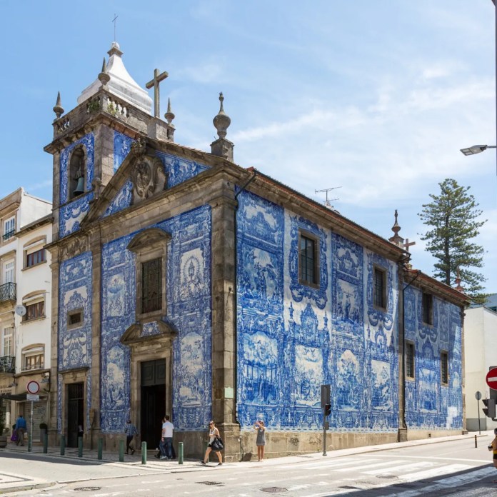 A full view of the Chapel of Souls in Porto, Portugal, covered entirely in 15,947 blue ceramic tiles (azulejos). The exterior features detailed religious scenes and ornate patterns created by Eduardo Leite in 1929. The church’s bell tower, cross, and two-story facade are prominent against a sunny sky. Pedestrians walk past the building, highlighting its grandeur and integration into the urban surroundings.