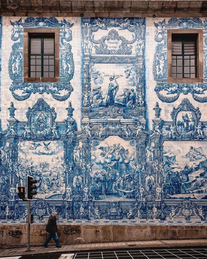 A section of the Chapel of Souls in Porto, Portugal, showcasing its iconic 15,947 blue ceramic tiles (azulejos). The intricate artwork depicts religious scenes, including Saint Francis of Assisi and Saint Catherine, framed by ornate decorative designs. The tiles, created by Eduardo Leite in 1929, blend seamlessly with the building's classic architectural style. The scene captures two large windows, a pedestrian walking below, and a traffic light, highlighting the juxtaposition of historic art and modern life.