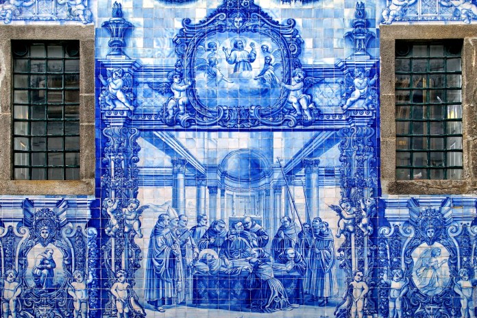 A detailed section of the Chapel of Souls in Porto, Portugal, showcasing its blue ceramic tiles (azulejos). The artwork illustrates a religious scene featuring figures gathered around a central figure in a domed architectural setting, flanked by decorative elements and angelic figures. The windows on either side frame the intricate design, blending the artistry of Eduardo Leite’s 1929 work with the church’s historical charm.