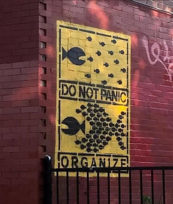 Mural on a red brick wall showing small black fish being chased individually in the top half, but grouped together to form a larger fish in the bottom half with the words “DO NOT PANIC” and “ORGANIZE.”
