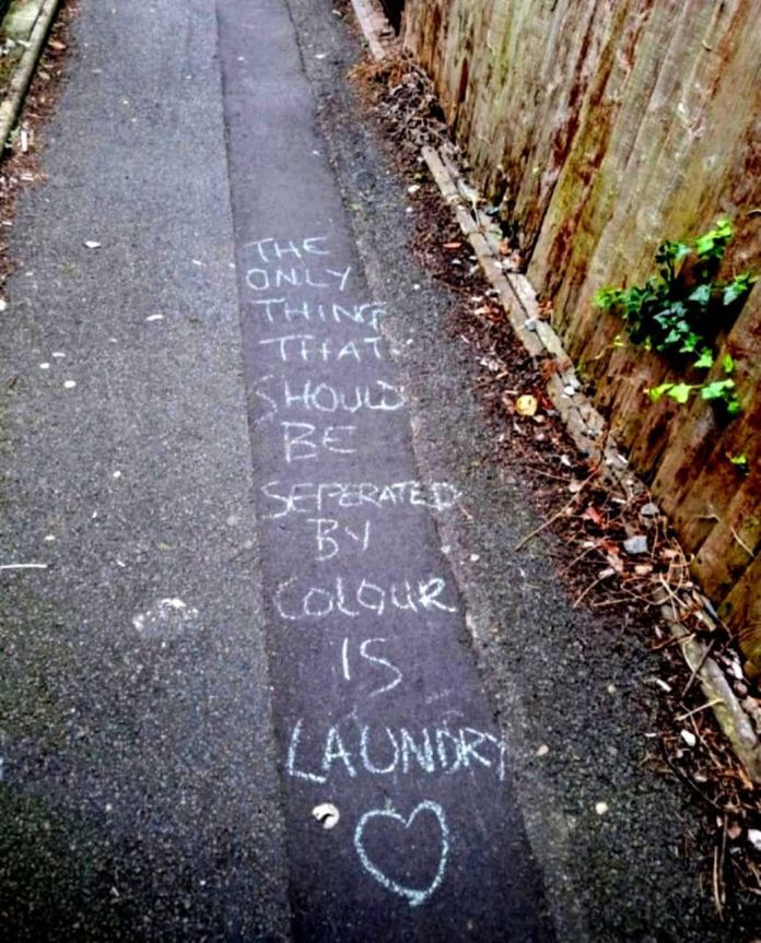 Chalk writing on a narrow pathway reads, “The only thing that should be separated by colour is laundry” with a heart symbol at the end.