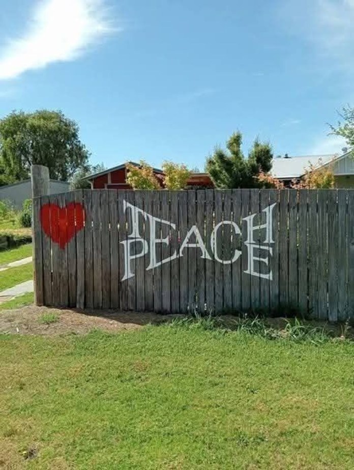 Fence in a grassy yard with a red heart painted next to large white letters that read “TEACH PEACE,” promoting nonviolence and unity.