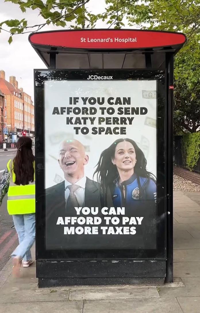Edited bus stop ad shows Jeff Bezos laughing with Katy Perry in a blue space suit. Caption reads: “If you can afford to send Katy Perry to space, you can afford to pay more taxes.” Background includes floating money, location tagged as St Leonard’s Hospital, London.