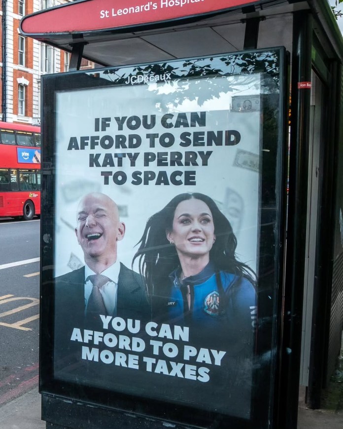 Edited bus stop ad shows Jeff Bezos laughing with Katy Perry in a blue space suit. Caption reads: “If you can afford to send Katy Perry to space, you can afford to pay more taxes.” Background includes floating money, location tagged as St Leonard’s Hospital, London.