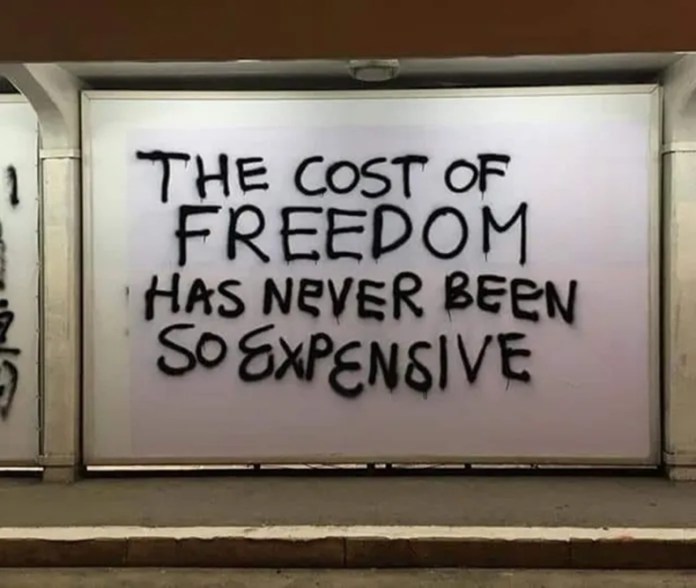 A white panel in a bus shelter features the black spray-painted phrase: “THE COST OF FREEDOM HAS NEVER BEEN SO EXPENSIVE.”
