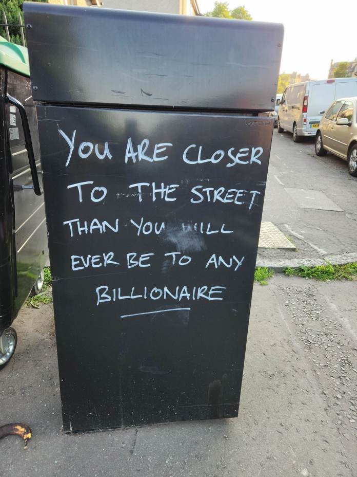 White handwritten text on a black discarded refrigerator reads: “YOU ARE CLOSER TO THE STREET THAN YOU WILL EVER BE TO ANY BILLIONAIRE.”