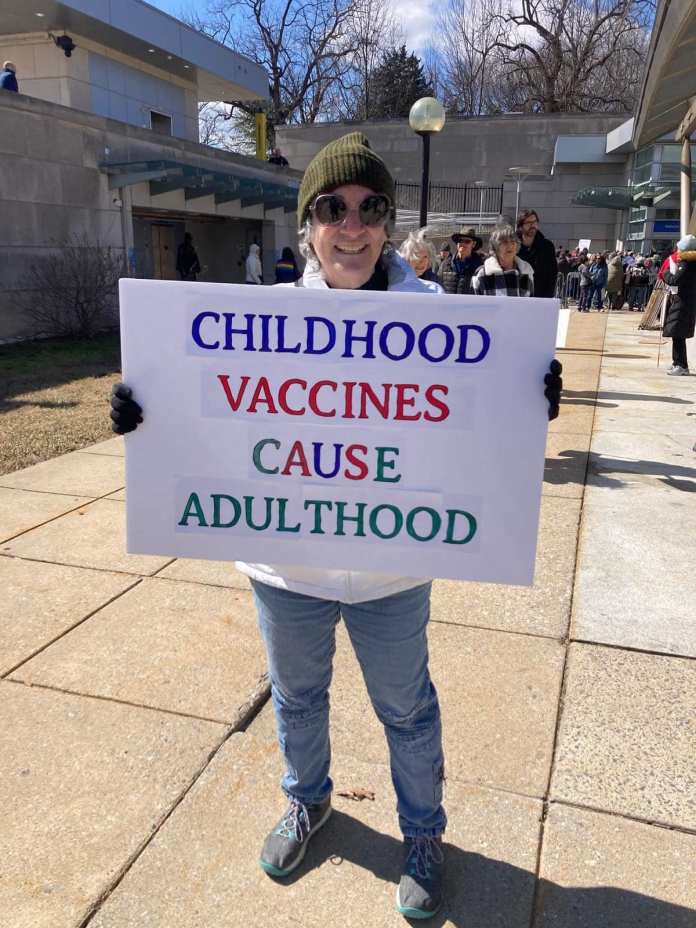 A protester holding a colorful sign reading "Childhood Vaccines Cause Adulthood" at a public protest.