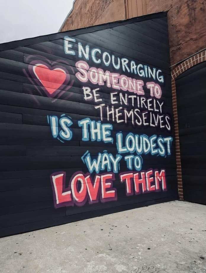 Mural reading "Encouraging someone to be entirely themselves is the loudest way to love them," painted brightly on black wood.