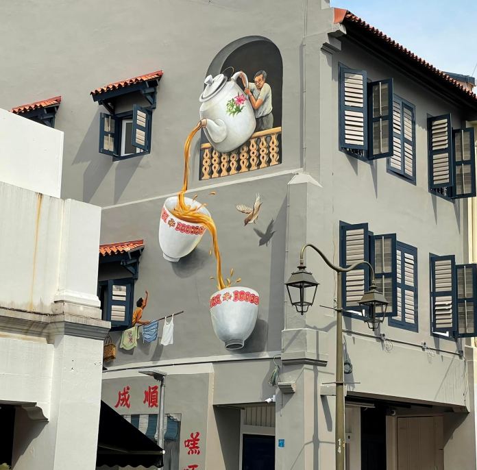 A 3D mural on the side of a building in Singapore’s Chinatown depicts a man pouring tea from a large white teapot into two oversized cups. The illusion includes a woman hanging laundry and a bird mid-flight, all integrated with the real structure's balconies and windows.