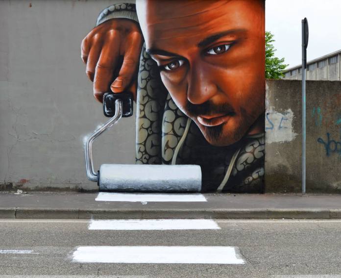 A mural on a street wall shows a hyper-realistic man holding a large paint roller, seemingly painting the white stripes of a crosswalk directly onto the road, creating a seamless visual illusion.