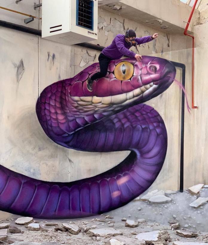 A massive purple viper with yellow eyes is painted to appear as though it’s slithering out of a broken wall. A person in a purple hoodie is seen climbing the painted snake, blending real and painted elements.