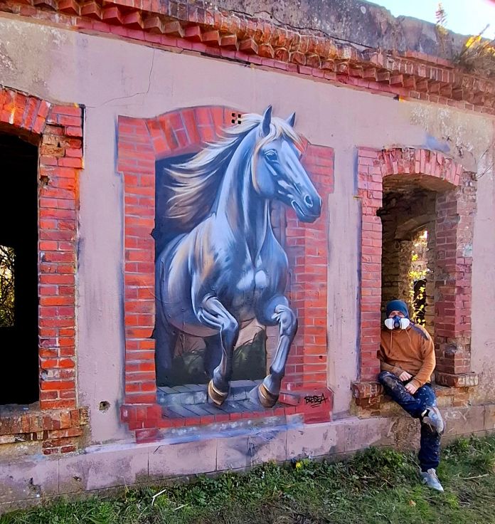 Mural by Blesea in Cherbourg, France, showing a powerful grey horse bursting out of a fake brick-framed opening on an abandoned building, with the artist sitting nearby in a face mask.