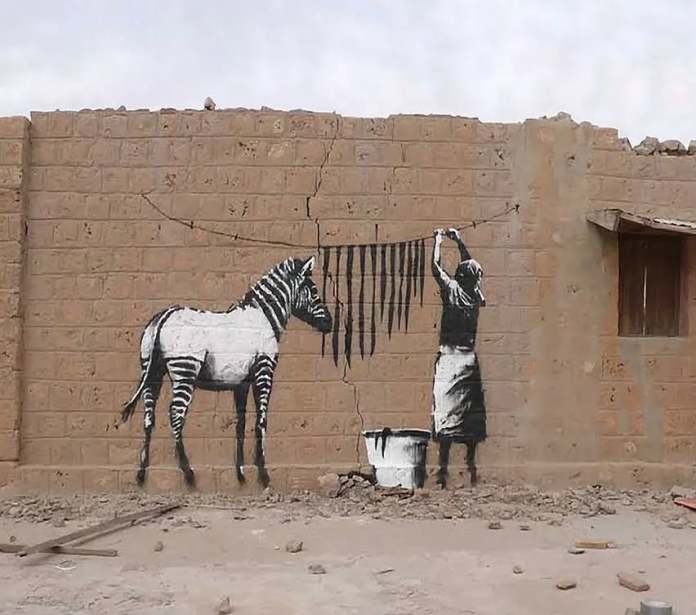 Street art by Banksy showing a stencil of a woman hanging black stripes on a clothesline, visually aligning with a white donkey to create the illusion of a zebra on a cracked brick wall.