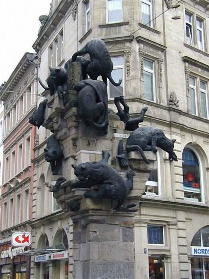 Column in Braunschweig, Germany, covered in dark bronze sculptures of cats in various poses—climbing, hanging, and crouching—on a stone base in a city center.