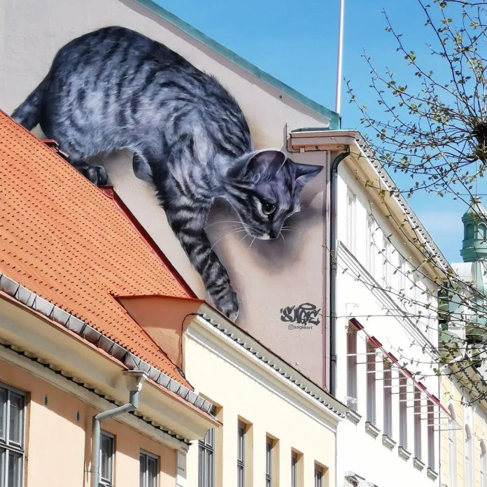 Giant mural of a grey tabby cat crouching on a rooftop edge in Kristianstad, Sweden, with lifelike fur detail and focused eyes, painted across a slanted wall.