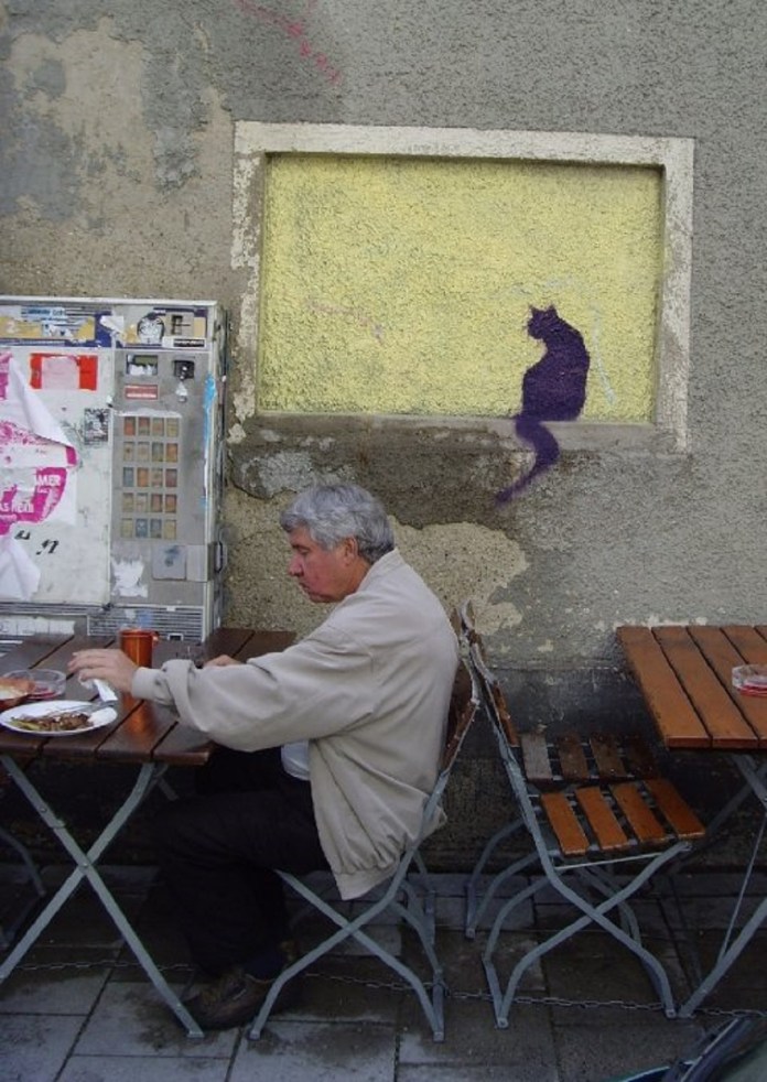 Black graffiti silhouette of a cat painted in a wall niche above a seated man at an outdoor table, blending into the urban background.