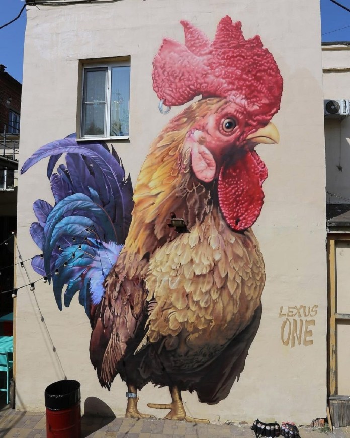 A hyperrealistic street art mural of a rooster with a red comb and vibrant blue-purple tail feathers, painted on a pale building wall in Russia, signed "LEXUS ONE."