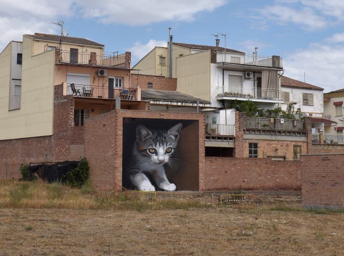 Mural of a lifelike gray tabby kitten crouched against a black background, painted on a red-brick wall among residential buildings in Spain.