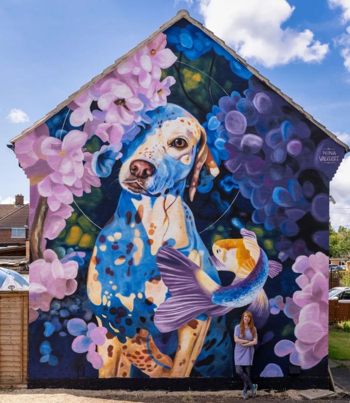 A vivid street art mural of a speckled dog surrounded by large flowers and a goldfish, painted in purple and blue tones across the gable of a UK house.