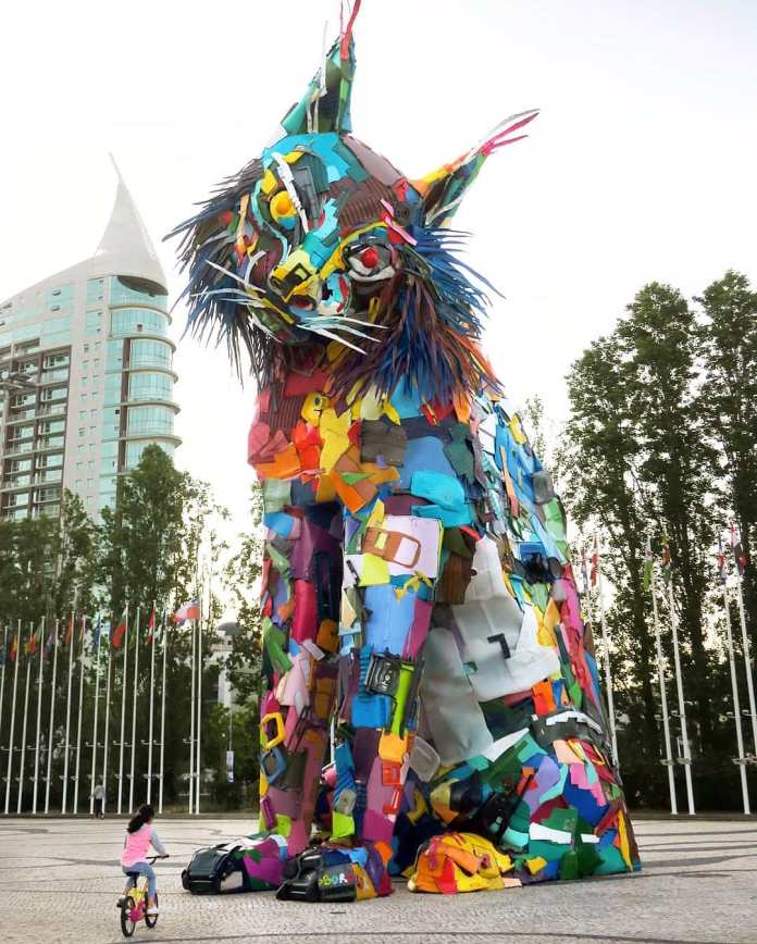 A towering lynx cat sculpture built from multicolored plastic and urban waste, placed in a modern plaza with flags and high-rises in the background.