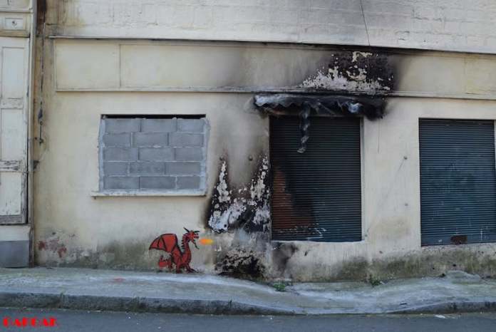 A small red painted dragon on a wall appears to breathe fire onto an actual burn-marked facade, blending art with real damage.