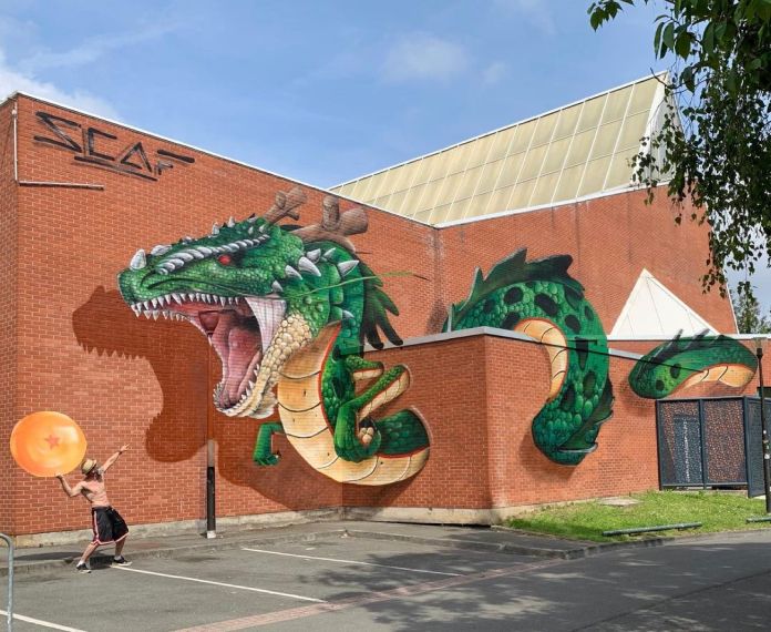 A 3D mural of Shenron by Scaf in Roubaix, France, painted on a red brick building to appear as if the green dragon is erupting from the wall; a person poses below holding a large orange Dragon Ball.
