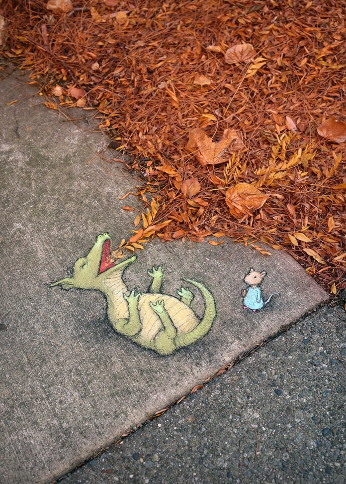 A sidewalk chalk drawing by David Zinn in Ann Arbor, Michigan, showing a green cartoon dragon on its back laughing while a small mouse in a dress looks on; real orange leaves surround the scene, blending with the art.