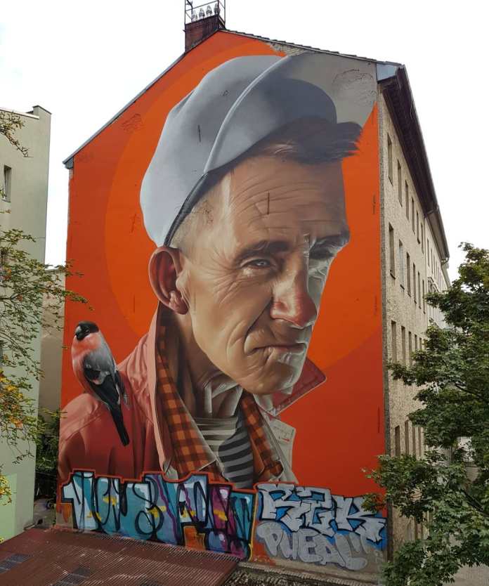 A striking mural by SMUG in Kreuzberg, Berlin, created for the Berlin Mural Fest. The artwork features a hyper-realistic portrait of an elderly man with weathered facial features, wearing a light gray cap, a red jacket with a checkered shirt underneath, and a black-and-white striped shirt beneath that. Perched on his shoulder is a detailed bullfinch bird with vibrant red and black plumage. The background is painted in a bold orange hue, contrasting with the lifelike details of the subject. At the bottom, colorful graffiti tags add an urban, layered aesthetic to the wall of this multi-story building.