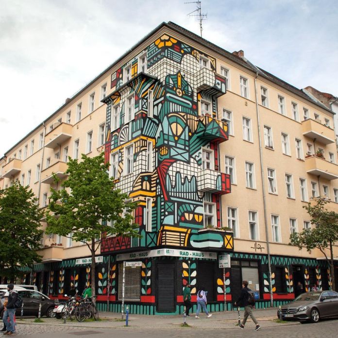 A vibrant street art mural by Isakov located at the intersection of Samariterstraße and Rigaer Straße in Friedrichshain, Berlin, Germany. The mural covers the corner of a multi-story residential building, featuring bold geometric patterns and colorful abstract designs in red, teal, yellow, black, and white. The design incorporates elements of urban and architectural motifs, blending seamlessly with the building's windows and balconies. The lower level, labeled "Rad-Haus," includes matching designs with foliage patterns, tying the entire facade together. Trees, pedestrians, and bicycles in the foreground enhance the lively urban setting.