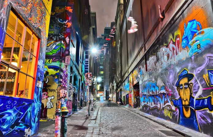 A vibrant night view of Hosier Lane in Melbourne, Australia, renowned for its ever-evolving street art and graffiti. The narrow cobblestone alley is illuminated by bright streetlights, casting a glow over the colorful murals that adorn both walls. On the left, a bright yellow-framed window spills warm light onto the street, while the right side showcases intricate graffiti, including a caricature of a man in a blue hat. Overhead, a string of shoes hangs from a wire, adding a whimsical touch to the urban landscape. The scene captures the dynamic energy and creativity of this iconic art-filled laneway.