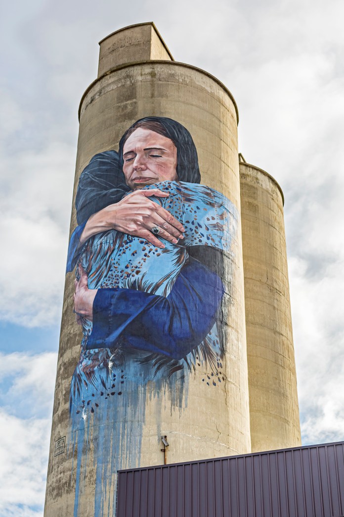 A powerful mural by Loretta Lizzio on a large concrete silo in Brunswick, Australia. The artwork depicts two figures embracing with deep emotion, highlighting themes of unity and compassion. One figure, wearing a dark headscarf, holds the other tightly, who is draped in a patterned blue garment. The mural is strikingly realistic, with subtle details such as the folds in the clothing and the expression of peace on their faces. The neutral tones of the concrete silo contrast with the vivid blues and blacks of the mural, making the piece stand out against the cloudy sky. Photo by Andrew.