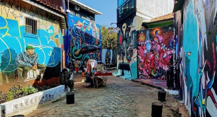 A colorful scene from Batman Alley (Beco do Batman) in São Paulo, Brazil, showcasing vibrant street art on both sides of a narrow cobblestone lane. The walls are adorned with diverse murals, including a realistic portrait of an elderly man in a green hat on the left and an explosion of abstract floral and cosmic designs on the right. In the center of the alley, a small table is set up with items for sale under a black umbrella, adding a lively and local market vibe to the artistic surroundings. The sunny day highlights the vivid colors and creative energy of this iconic destination.