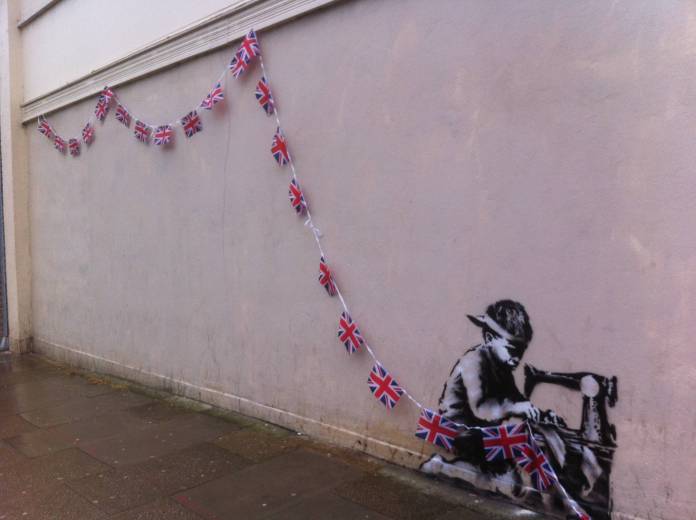 A street art piece by Banksy from 2012 in London, created to mark the Queen’s Diamond Jubilee. The artwork features a stencil of a young boy sitting at a sewing machine, crafting Union Jack bunting. The realistic details of the boy’s concentrated expression contrast sharply with the whimsical nature of the celebratory flags, which extend in a line along the wall. The placement on a plain beige wall adds to the stark and thought-provoking commentary on labor and festivity. The scene reflects Banksy’s signature style of blending humor with social critique.