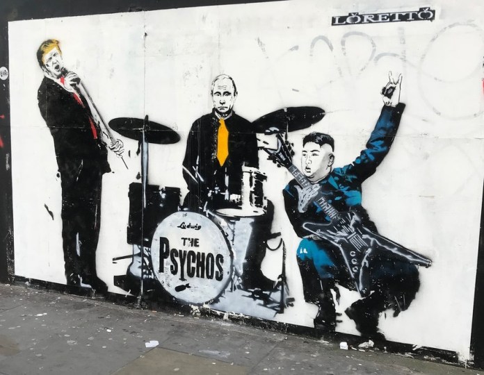 A provocative mural titled "The Psychos" by artist Loretto, painted in 2018 in Shoreditch, London, UK. The artwork portrays three world leaders as members of a fictional band. Donald Trump is depicted as the vocalist, holding a microphone, Vladimir Putin sits at a drum set with a stern expression, and Kim Jong-un plays an electric guitar, striking a flamboyant pose with a devil horns hand gesture. The drum set is labeled "The Psychos," highlighting the satirical theme. The black-and-white stencil style is accented by bright splashes of red, yellow, and blue in their ties and outfits, emphasizing their personalities. The piece critiques global power dynamics with sharp humor.