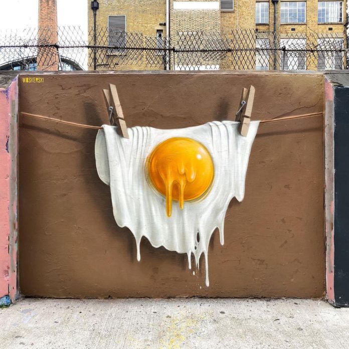A creative mural titled "Free Range Eggxaggeration" by WOSKerski, located in Shoreditch. The artwork depicts a hyper-realistic image of a white t-shirt hanging on a clothesline, with an oversized egg yolk melting and dripping down the fabric as if the shirt is made of egg. The rich yellow of the yolk contrasts strikingly with the brown wall background, making the surreal composition pop. The surrounding urban setting, with a wire fence and brick buildings in the background, enhances the playful and imaginative tone of the piece. The illusionistic style demonstrates the artist's mastery of depth and texture.