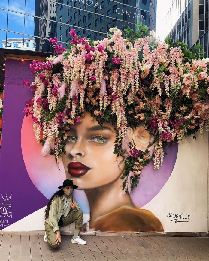 A stunning mural by OG Millie in New York, USA, depicting a stylized portrait of a woman with vibrant red lips, green eyes, and glowing skin, set against a soft gradient background of purples and pinks. The mural is elevated by a three-dimensional floral installation above the woman's head, featuring cascading pink, white, and purple flowers, along with lush green foliage, creating the appearance of a lavish floral crown. A person wearing a hat and face mask poses in front of the mural, adding a modern and interactive touch to the artwork's urban setting. Skyscrapers in the background reflect the bustling energy of the city.