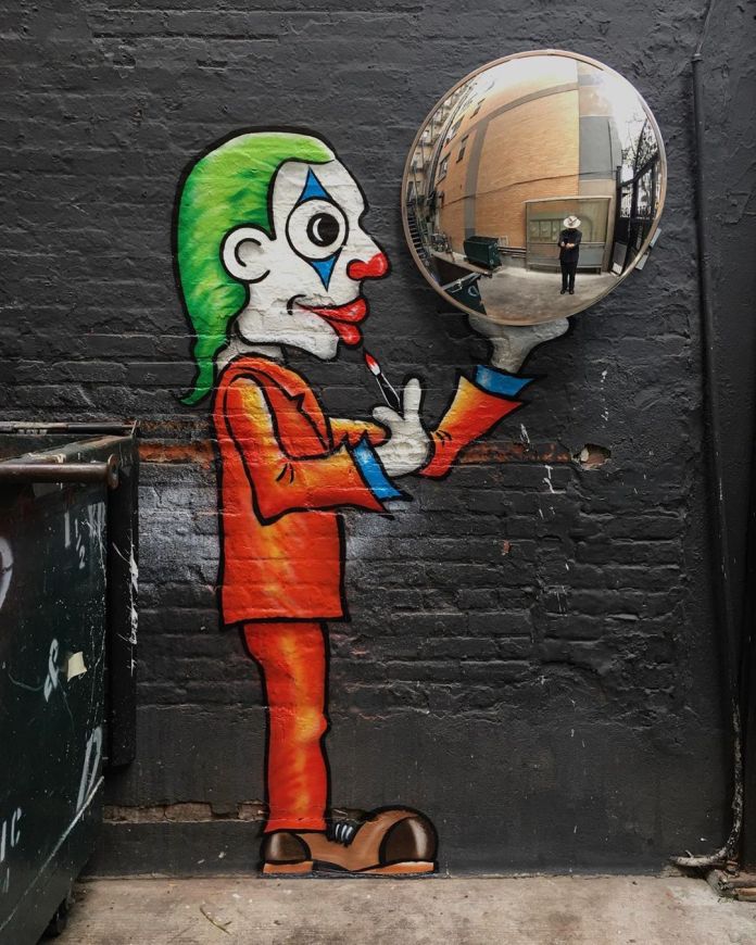 A playful mural by Tom Bob in New York, USA, featuring a cartoonish figure with green hair and clown makeup, dressed in an orange suit. The character is depicted applying red lipstick while holding a reflective circular mirror, integrated cleverly into the artwork. The mirror reflects a real-life scene of an alleyway, including a person in the background, blending the painted mural with its environment. The dark brick wall background and the adjacent dumpster add an urban, gritty context that contrasts with the vibrant and humorous style of the art.