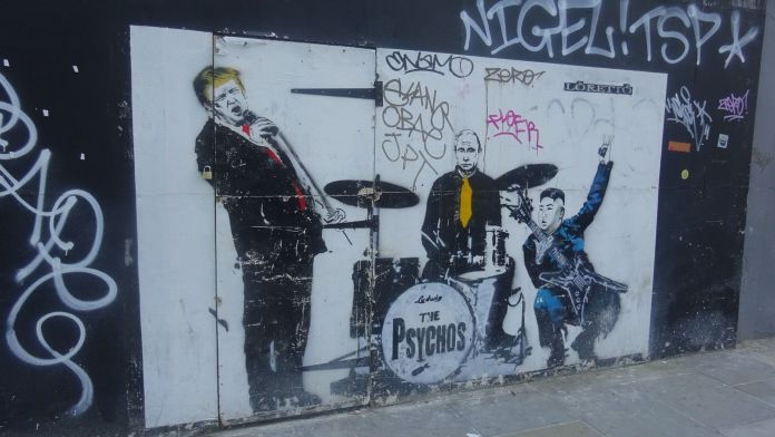 Satirical street art by Loretto from 2018 in Shoreditch, London, UK, depicting a mock band called 'The Psychos.' The mural features caricatures of world leaders: Donald Trump on vocals, Vladimir Putin on drums, and Kim Jong-un playing a guitar. Trump leans into the microphone, Putin sits stoically with a yellow tie, and Kim strikes a rockstar pose with a peace sign and an electric guitar. The band's name is boldly painted on the drum kit, adding a comedic yet politically charged edge to the artwork. Graffiti tags and a worn urban wall form the backdrop.