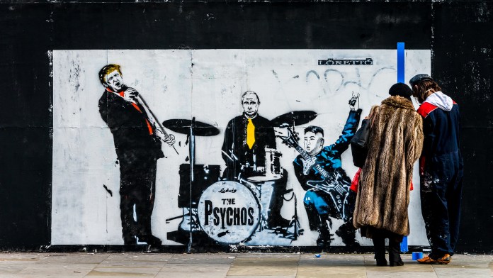 Satirical street art by Loretto from 2018 in Shoreditch, London, UK, depicting a mock band called 'The Psychos.' The mural features caricatures of world leaders: Donald Trump on vocals, Vladimir Putin on drums, and Kim Jong-un playing a guitar. Trump leans into the microphone, Putin sits stoically with a yellow tie, and Kim strikes a rockstar pose with a peace sign and an electric guitar. The band's name is boldly painted on the drum kit, adding a comedic yet politically charged edge to the artwork. Graffiti tags and a worn urban wall form the backdrop.