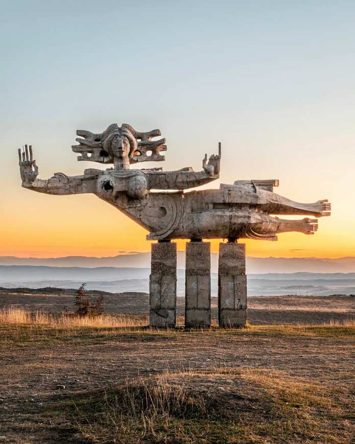Abstract concrete sculpture of a large winged female figure with open hands, standing on columns at sunset in the Georgian countryside.
