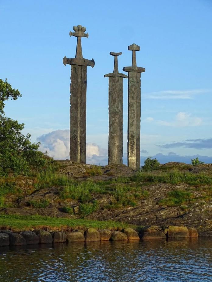 Three towering bronze Viking swords planted in a rocky hill beside a fjord, under a clear blue sky in Stavanger, Norway.