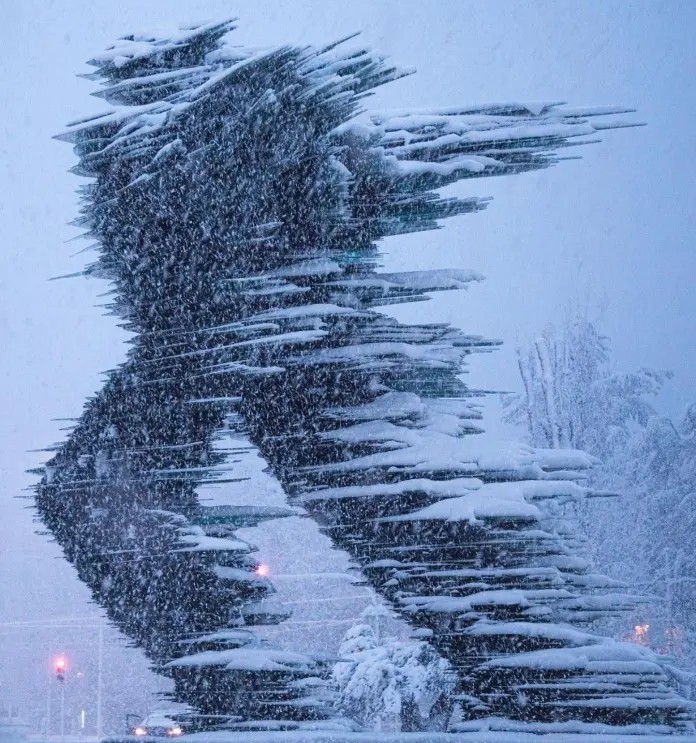 Glass sculpture of a running human figure composed of hundreds of sharp horizontal panes, covered in snow and standing in a snowy urban setting in Athens, Greece.