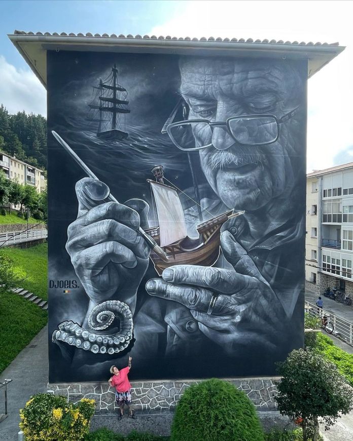 Monochrome mural of an elderly man crafting a ship model with ghost ships behind him and a curled tentacle below, painted on a large building wall.