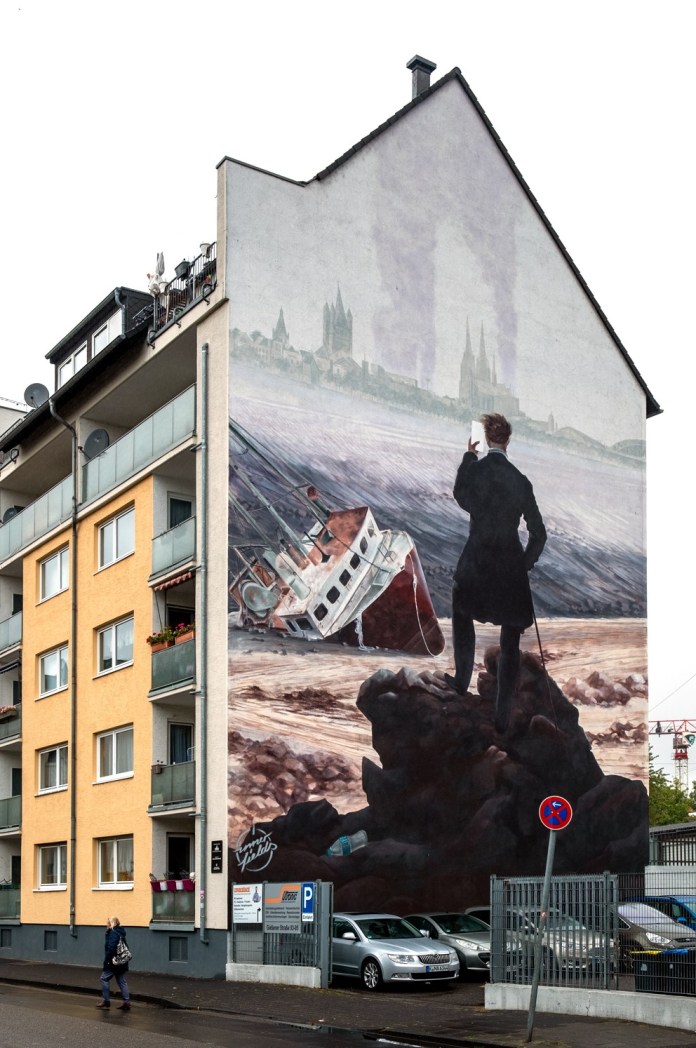 Large-scale mural of a man in black looking at a shipwreck and a misty city skyline, inspired by romantic landscape paintings, on a building facade.