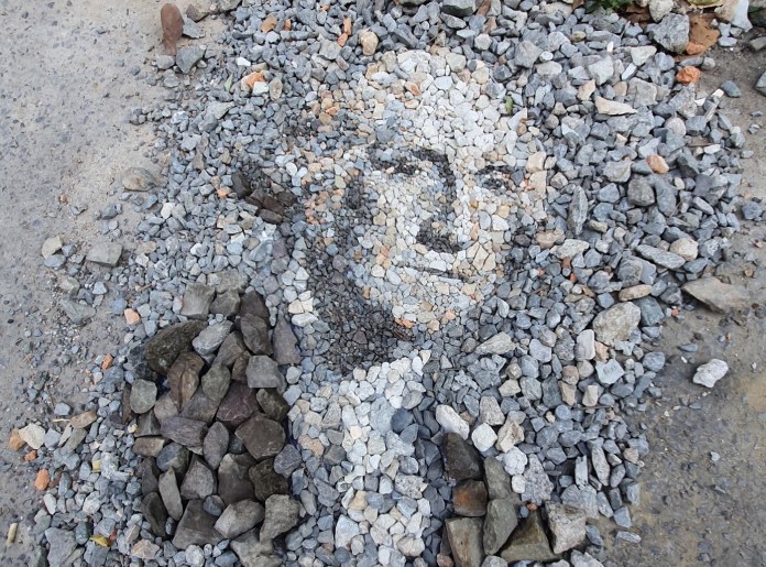A stone and pebble portrait of George Washington titled "George Washingstone," created by Justin Bateman in Bangkok, Thailand. The artwork uses carefully arranged stones of various sizes and colors to form the recognizable face of Washington, blending earthy tones to capture intricate details. The piece sits outdoors, surrounded by loose gravel, reflecting the ephemeral nature of Bateman's creations.