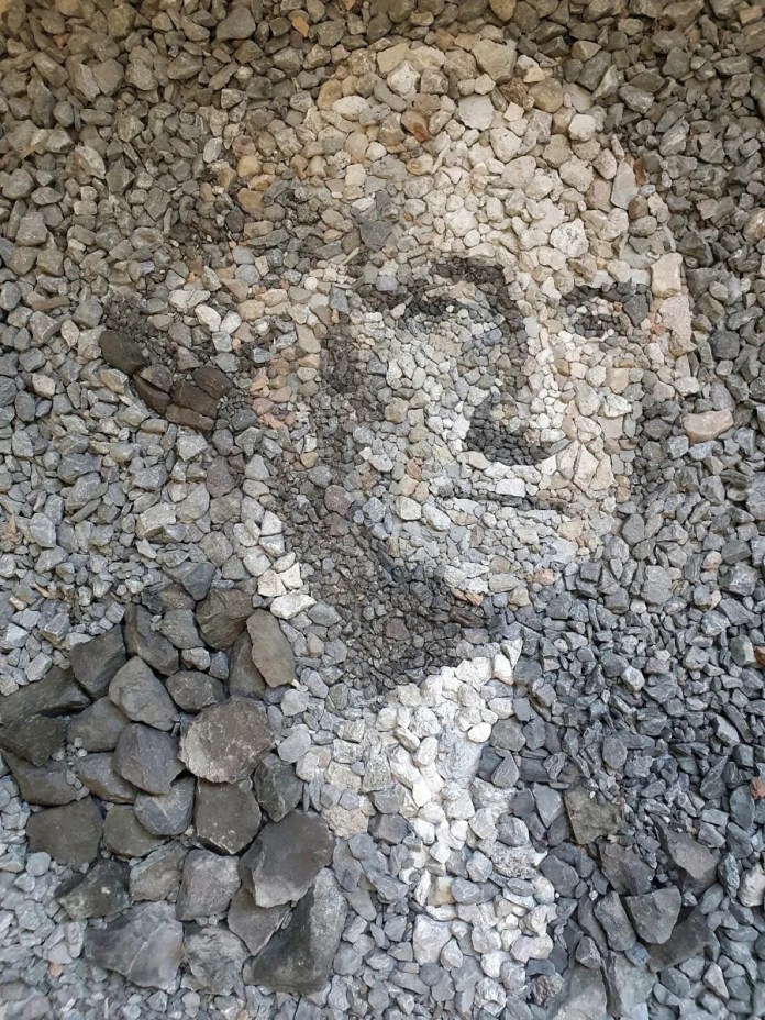 A detailed stone and pebble portrait of George Washington, titled "George Washingstone," created by Justin Bateman in Bangkok, Thailand. The arrangement uses varying shades of gray, white, and brown stones to bring out the contours and expressions of Washington’s face. The portrait sits in a defined area of gravel, showcasing Bateman's ability to use natural materials to recreate iconic imagery with precision.