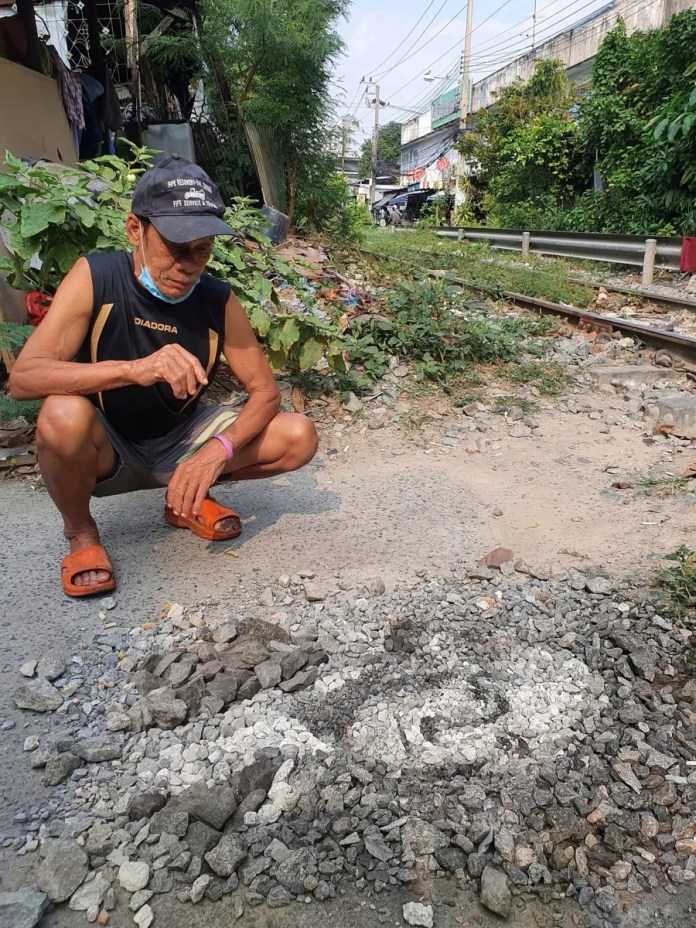 An elderly man crouches beside Justin Bateman's ephemeral pebble portrait of George Washington, titled "George Washingstone," created in Bangkok, Thailand. The man points towards the artwork on the ground, situated near a railway track surrounded by greenery and urban elements. The portrait blends seamlessly into the gravelly surface, highlighting its temporary and naturalistic essence.