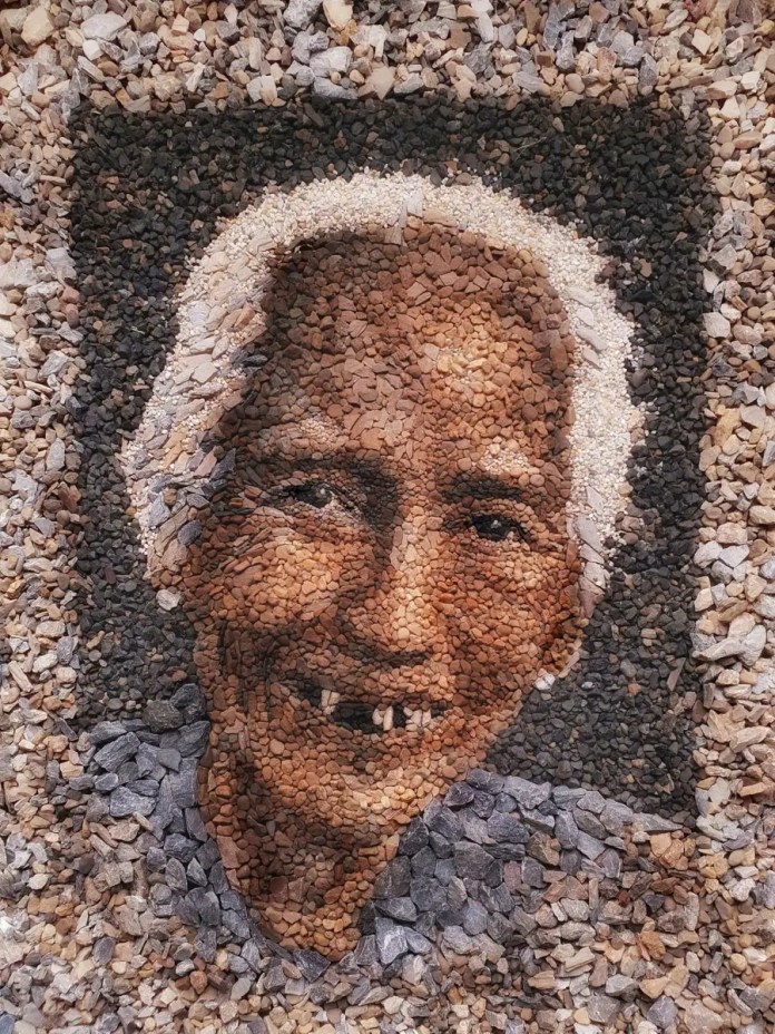 A stunning stone and pebble portrait titled "Grace" by Justin Bateman, depicting an elderly woman from Myanmar with a warm smile. The intricate arrangement of stones captures the depth of her facial features, using earthy tones and highlights to convey both strength and serenity. The portrait is framed within a border of darker stones, adding contrast and enhancing the subject’s prominence. Created in Thailand, this artwork reflects Bateman's skill in combining natural materials with emotional storytelling.